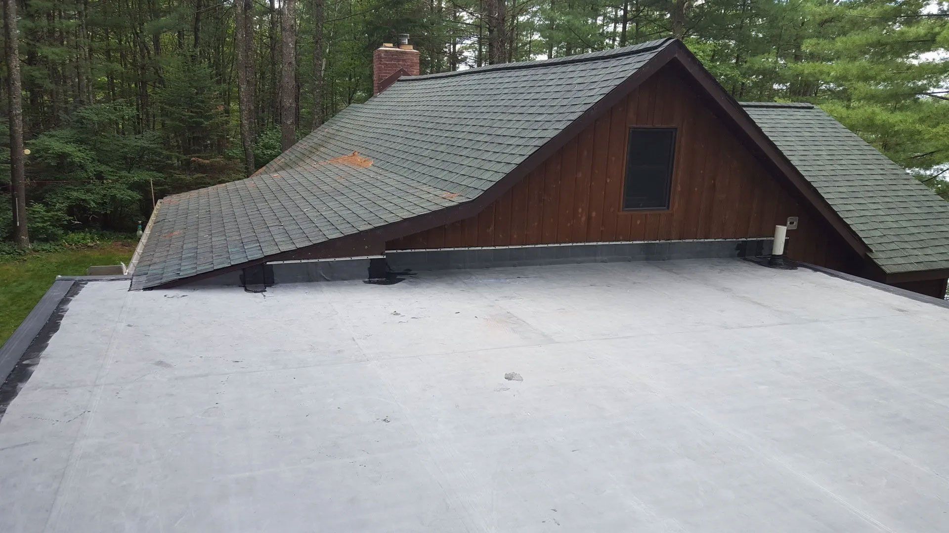 Flat white roof in foreground with a brown cabin roof and wooded background