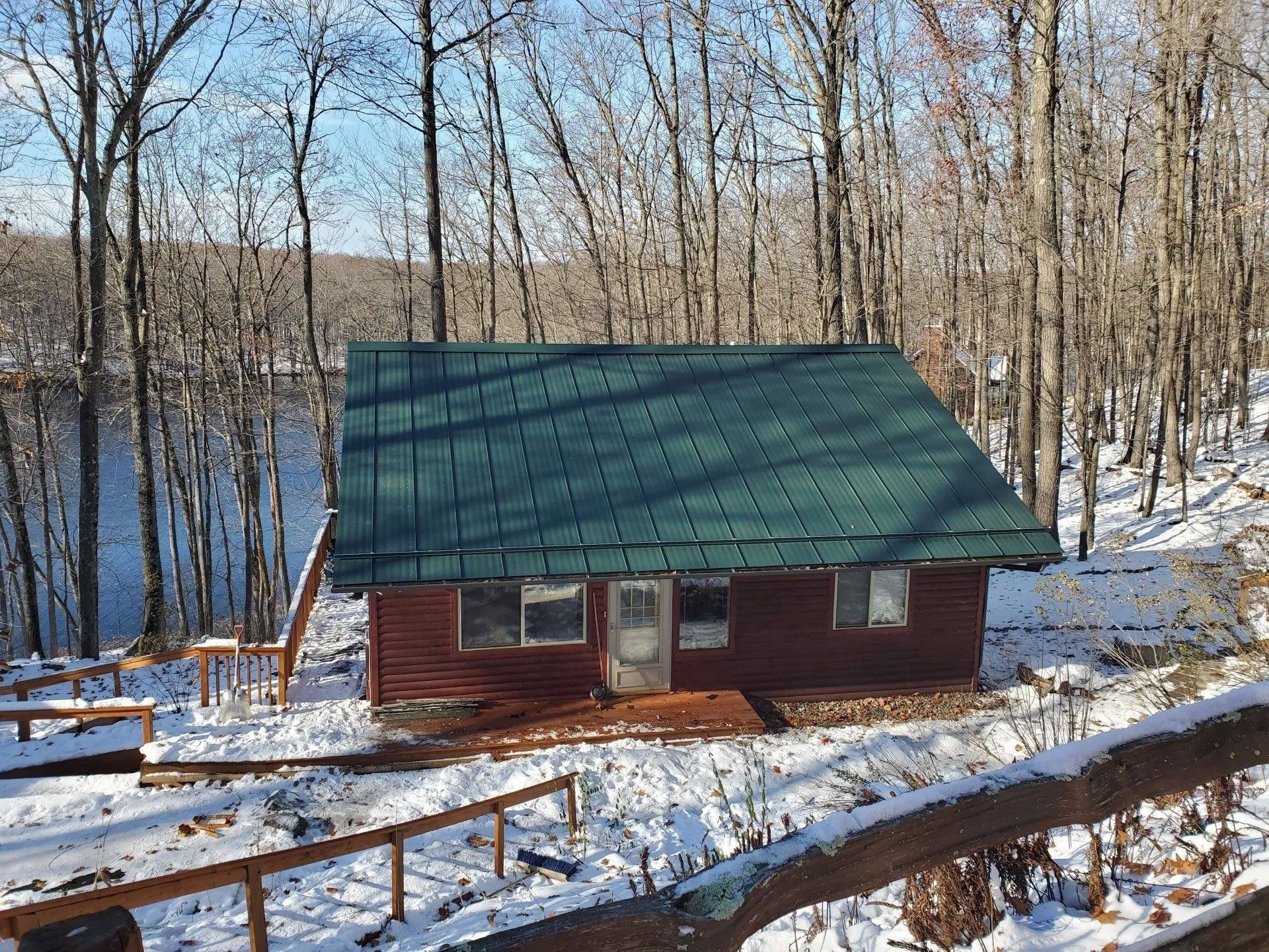 Small red cabin with green roof by a snowy lakeside in winter, surrounded by bare trees.