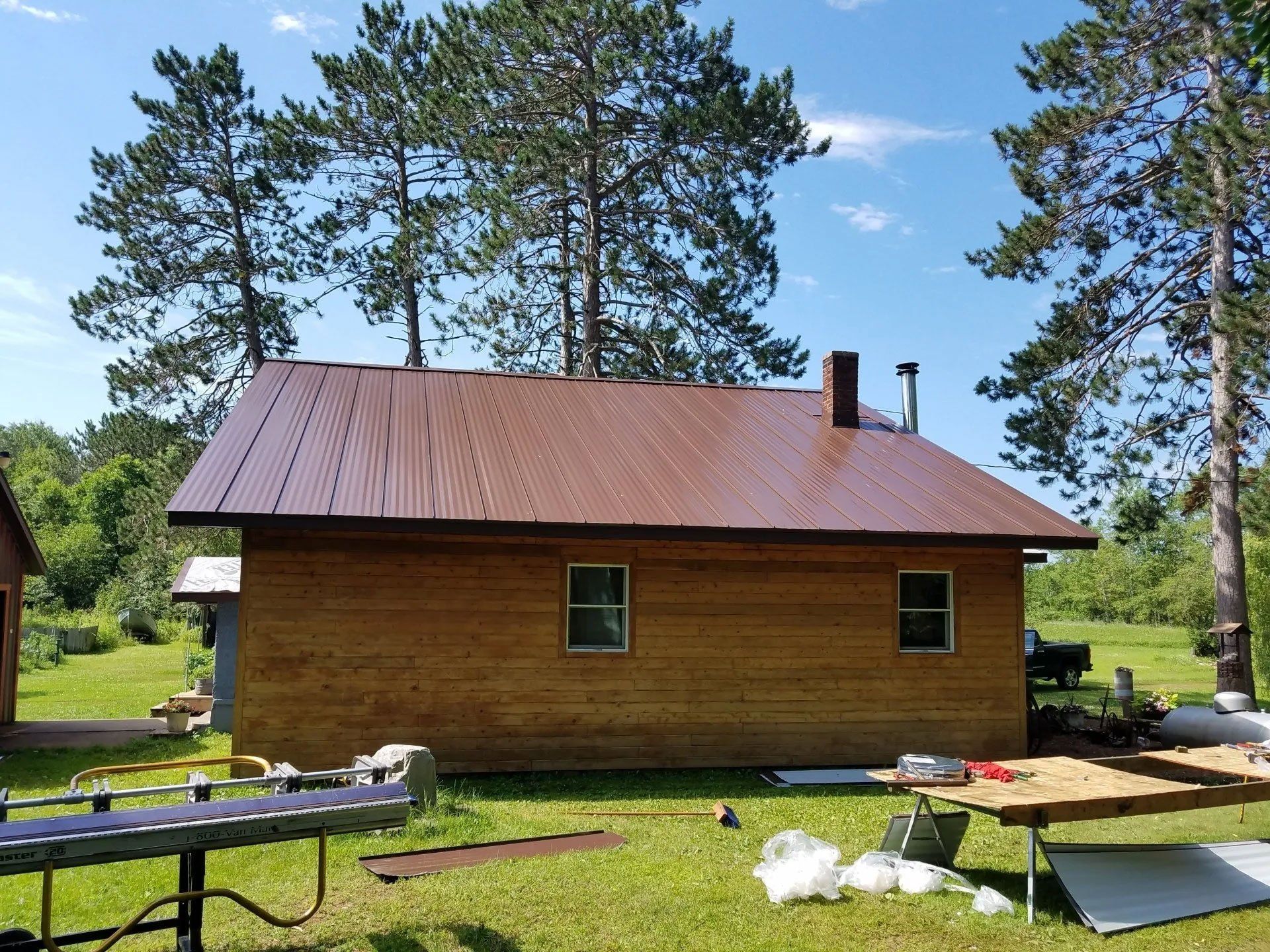 Wooden cabin with a brown metal roof on a sunny day, surrounded by trees and yard materials
