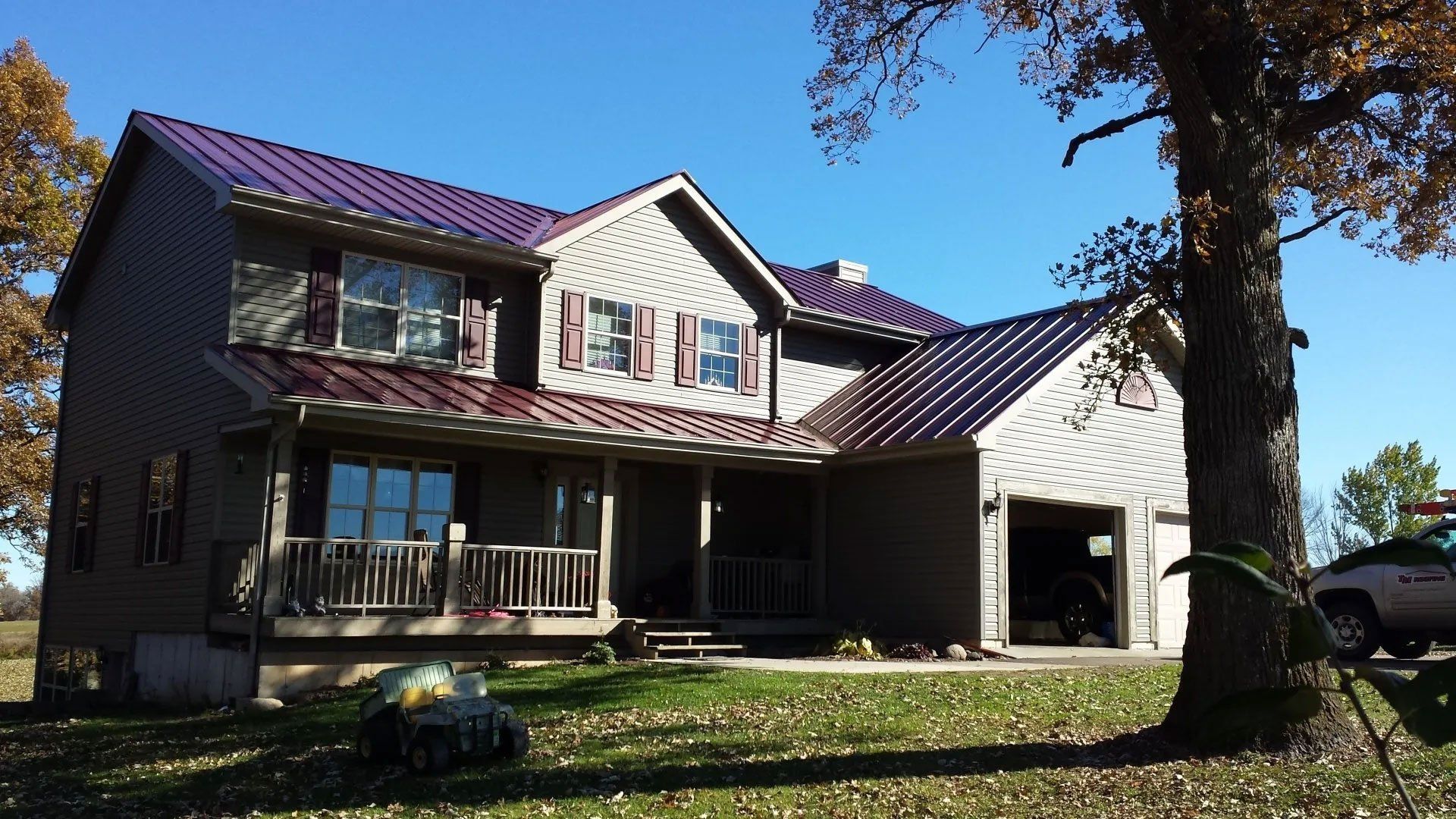 Two-story house with a covered porch, metal roof, and large trees in a sunny yard