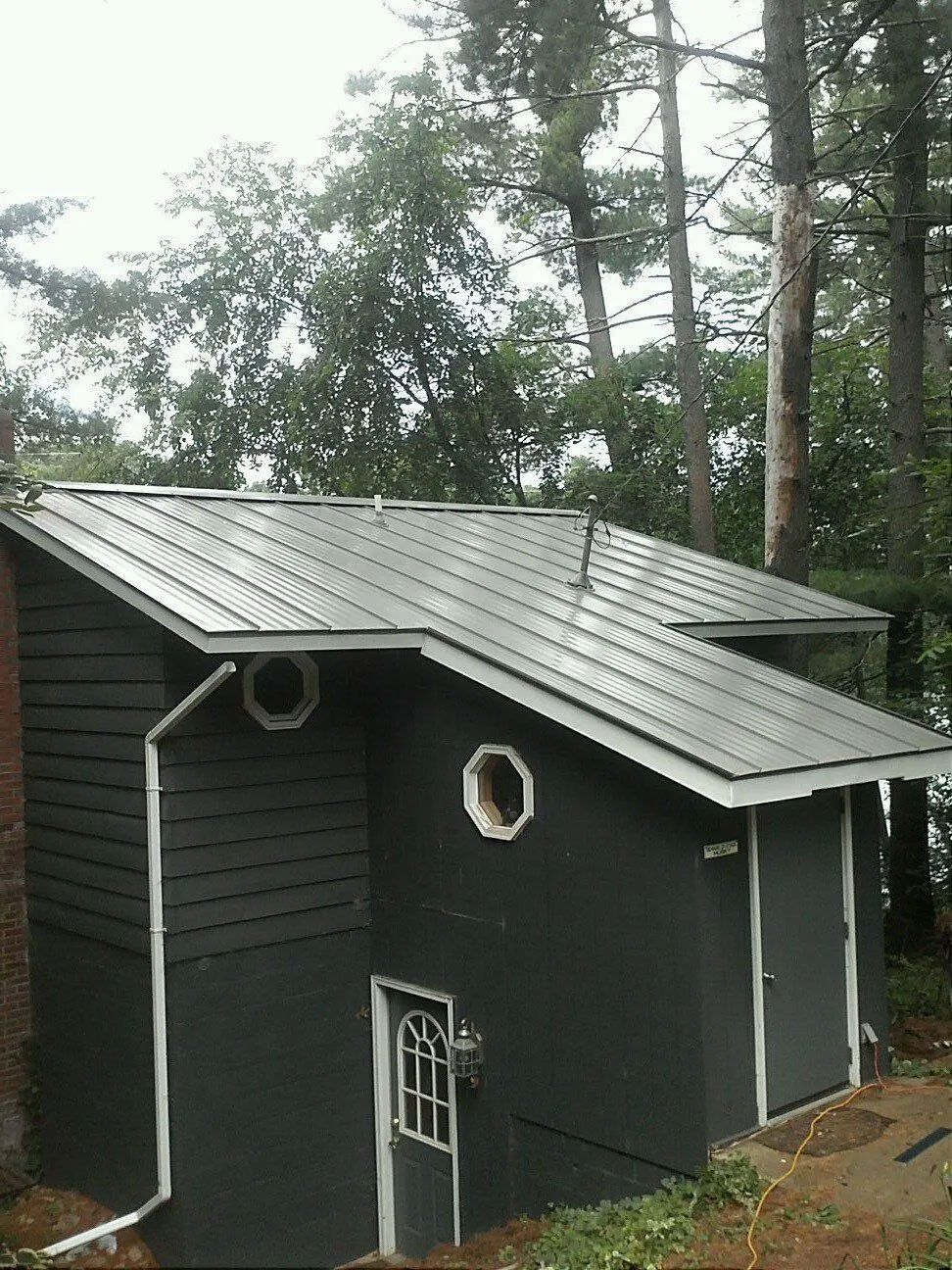 Small dark gray cabin with a metal roof in a wooded area, viewed from the side.