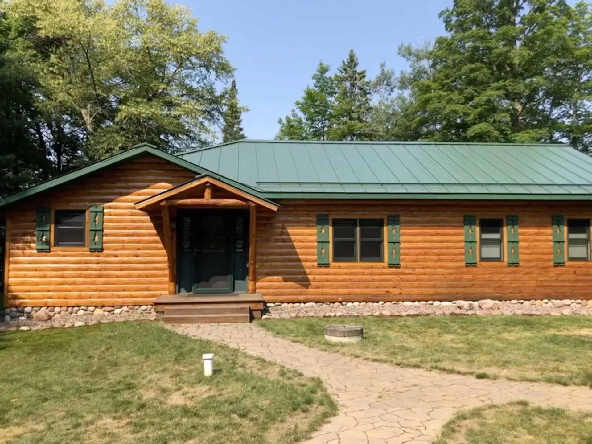 Wooden cabin with green metal roof and front porch, set in a grassy yard with a stone path