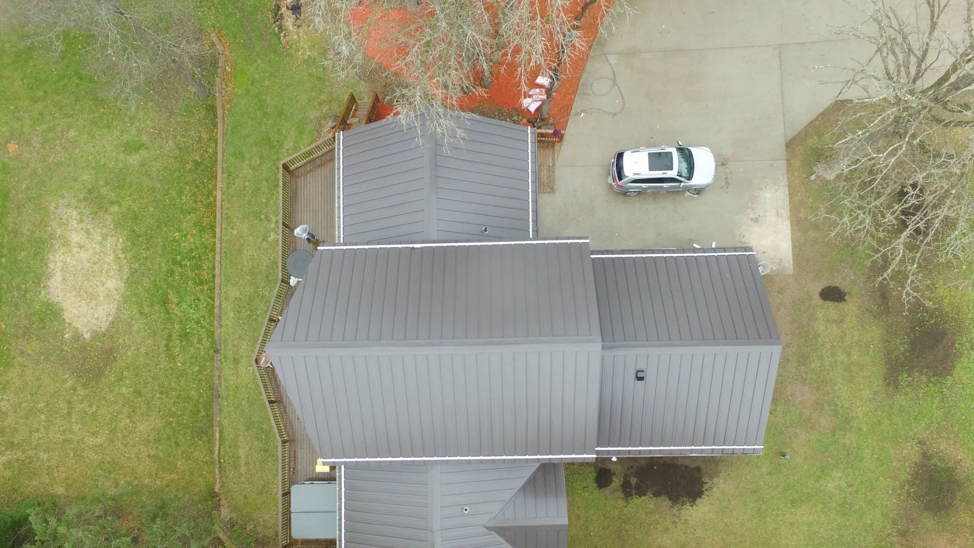 Aerial view of a house with gray roof beside a white car on a driveway, surrounded by green yard.