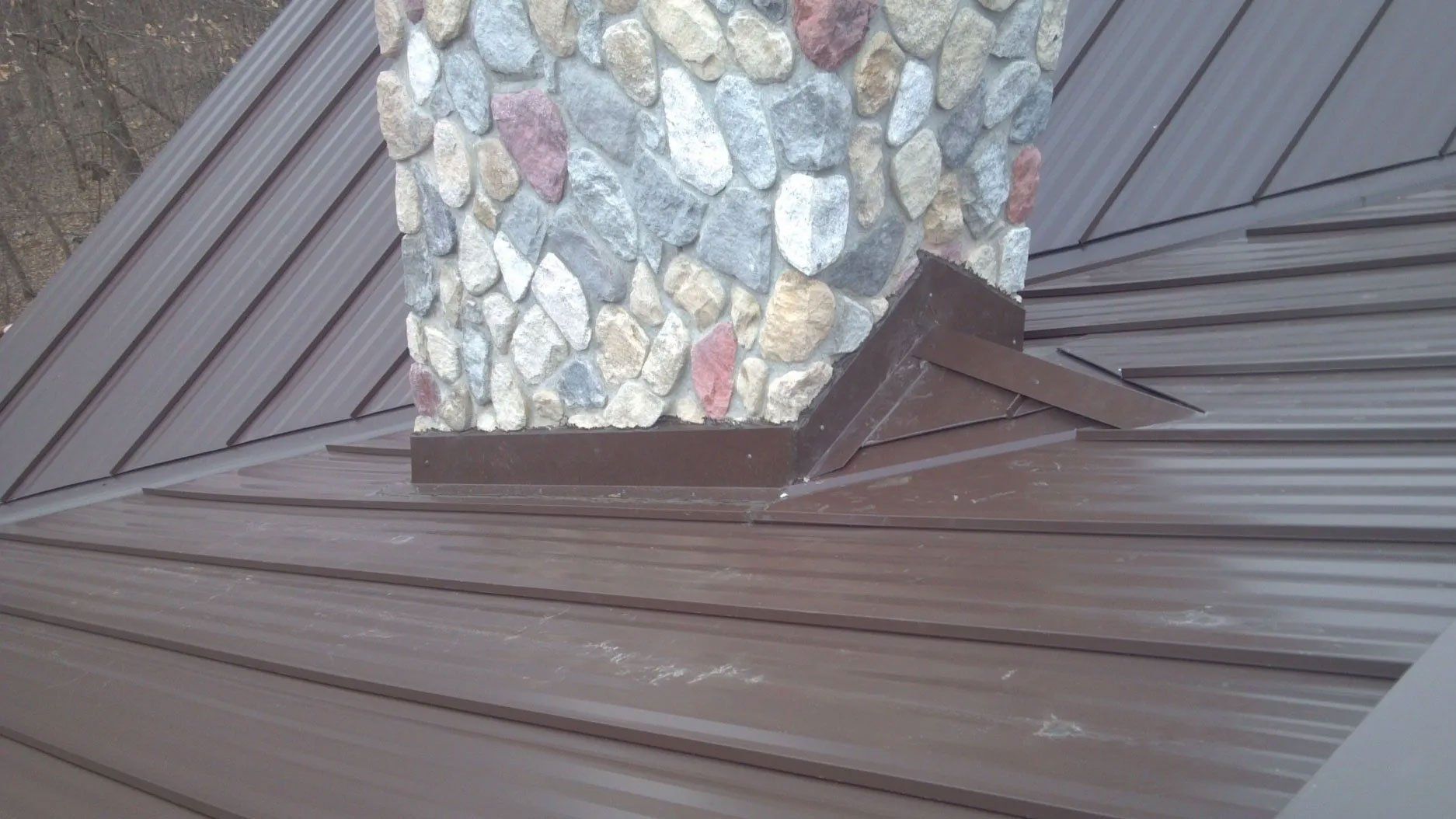 Brown metal roof flashing around a stone chimney with intersecting roof seams