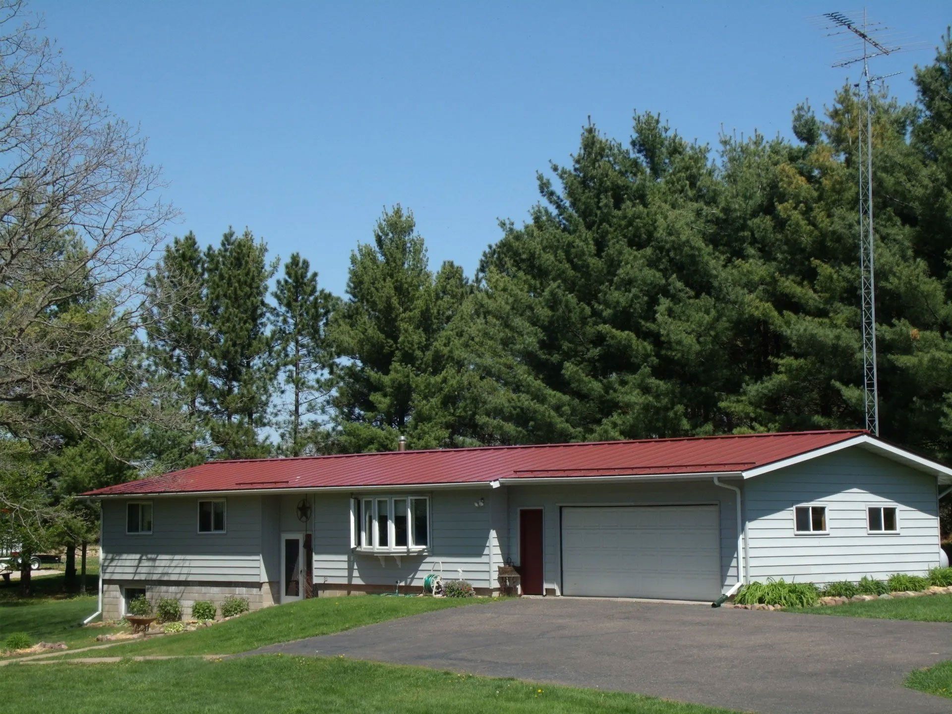 Single-story gray house with red roof, white garage, and driveway surrounded by trees under a blue sky