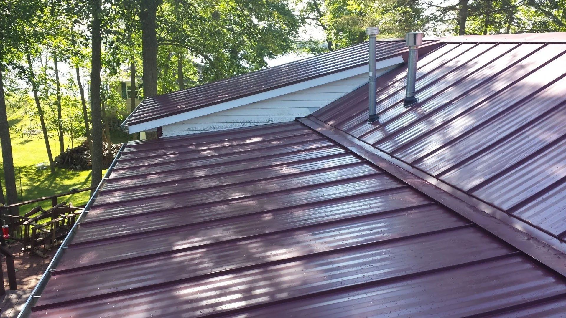 Metal roof with skylight and chimney pipes beside leafy trees in sunlight