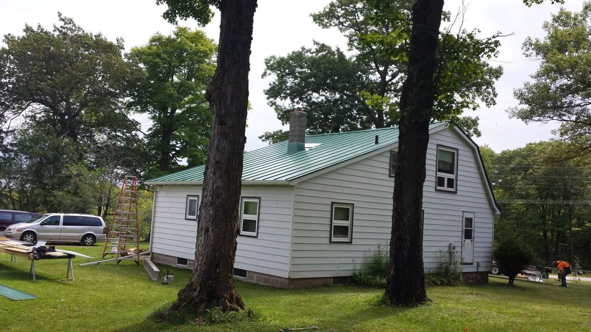 Small white house with a green metal roof in a grassy yard, framed by tall trees.