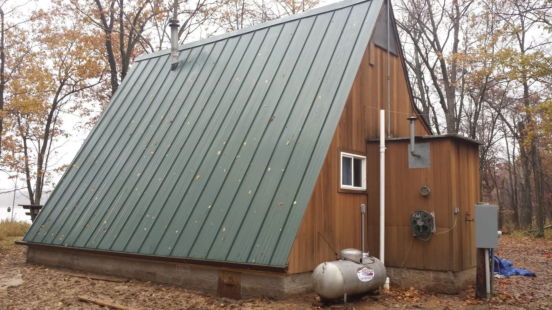 Small A-frame cabin with green metal roof and propane tanks in a wooded autumn setting