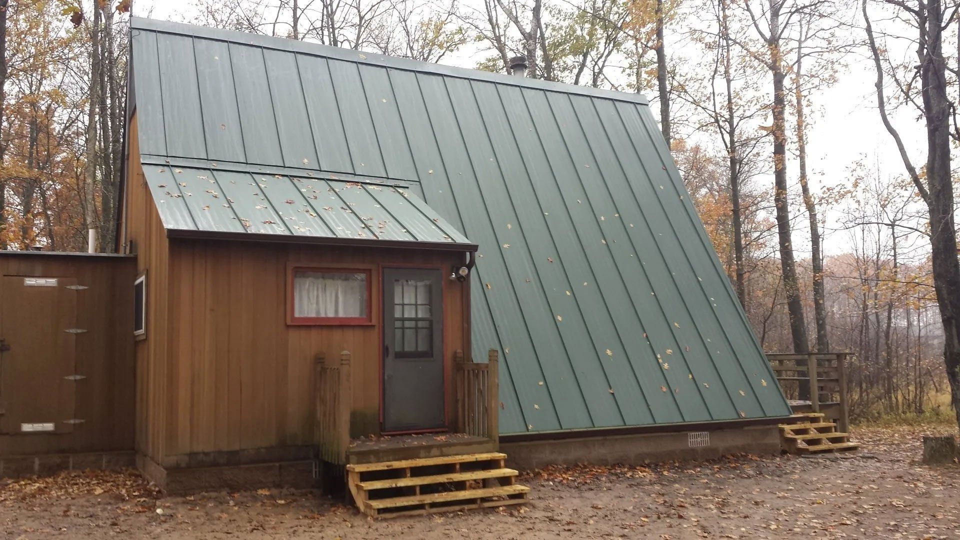 Small A-frame cabin with green metal roof and propane tanks in a wooded autumn setting
