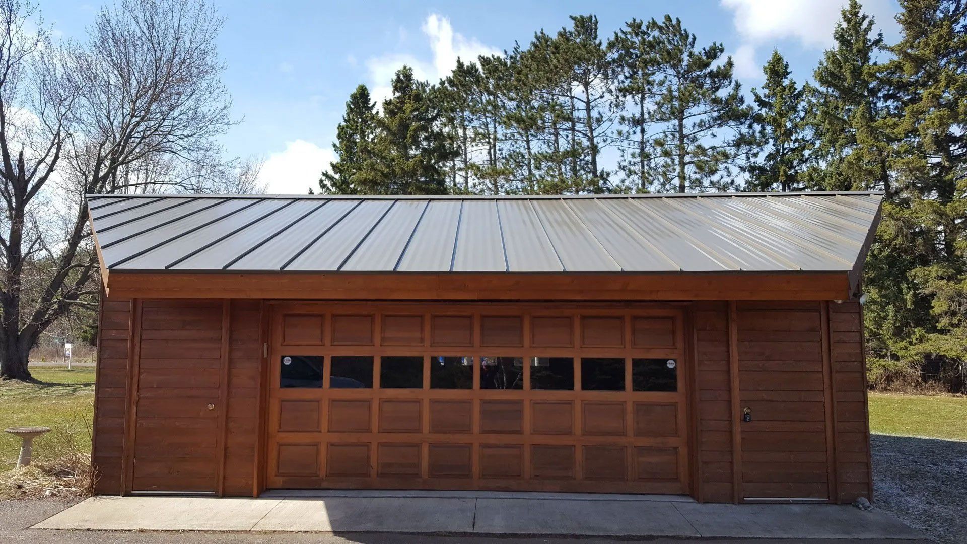 Small brown wooden garage with a gray metal roof and large front doors, set outdoors among trees.