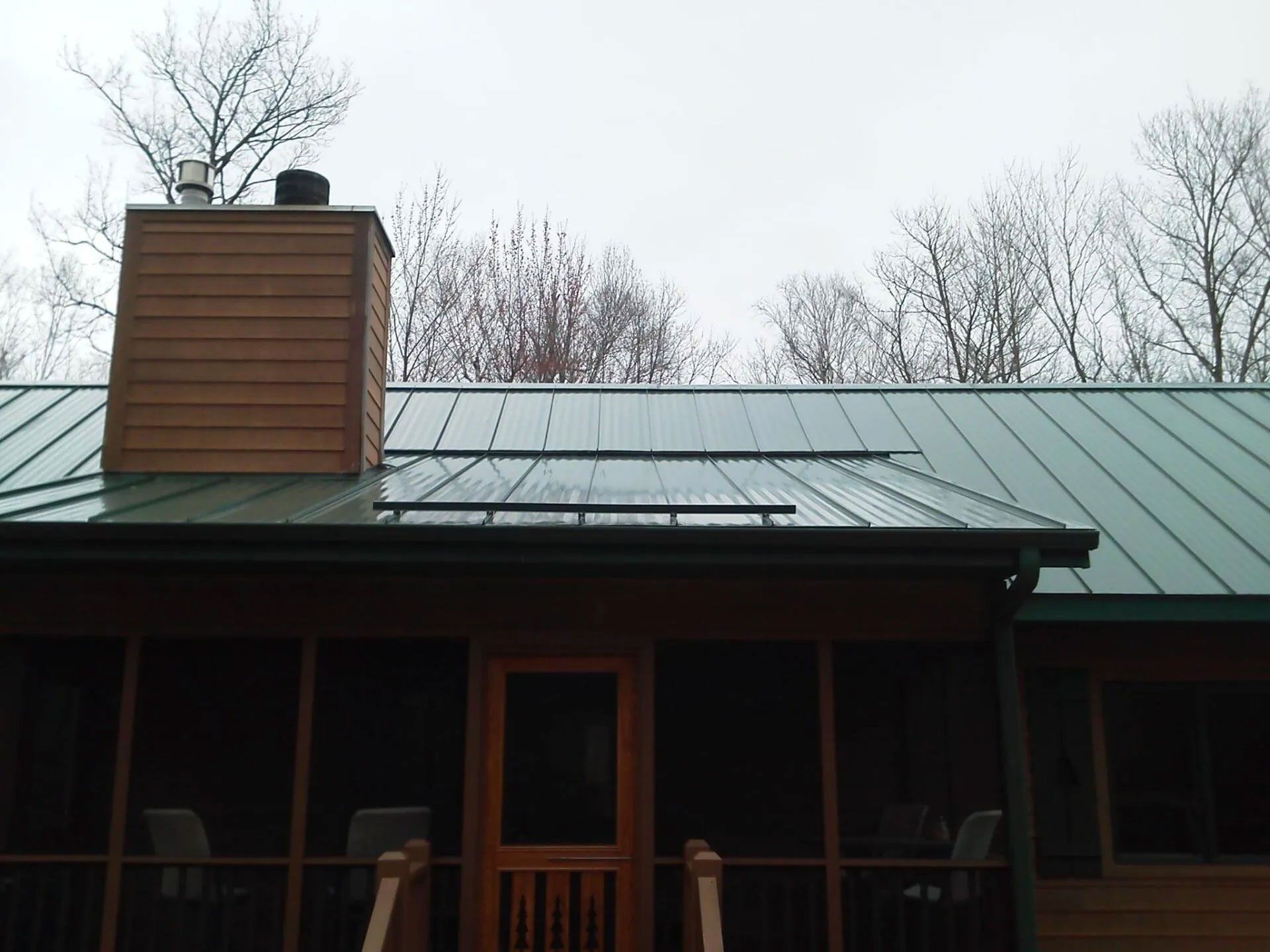 House with a metal roof and chimney under overcast sky, framed by bare trees