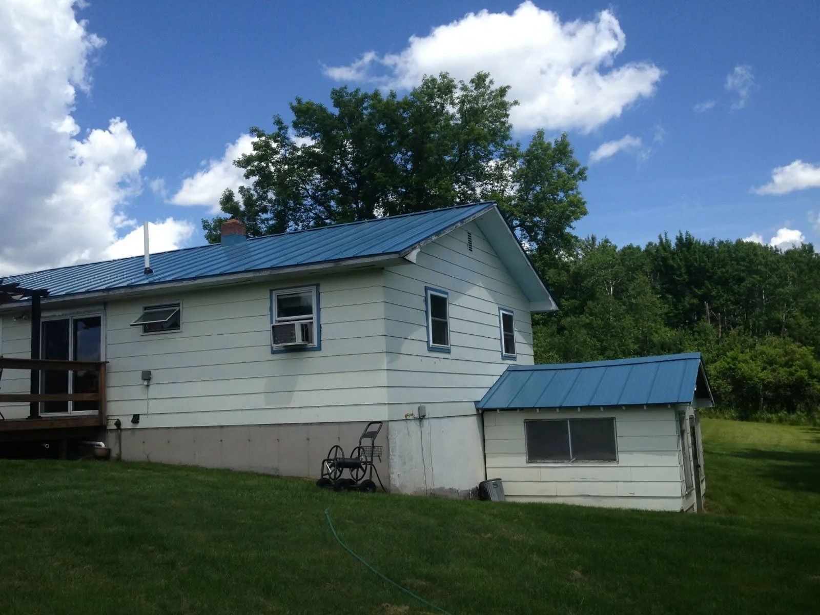 White house with blue metal roof and small shed on a grassy hillside under a cloudy sky
