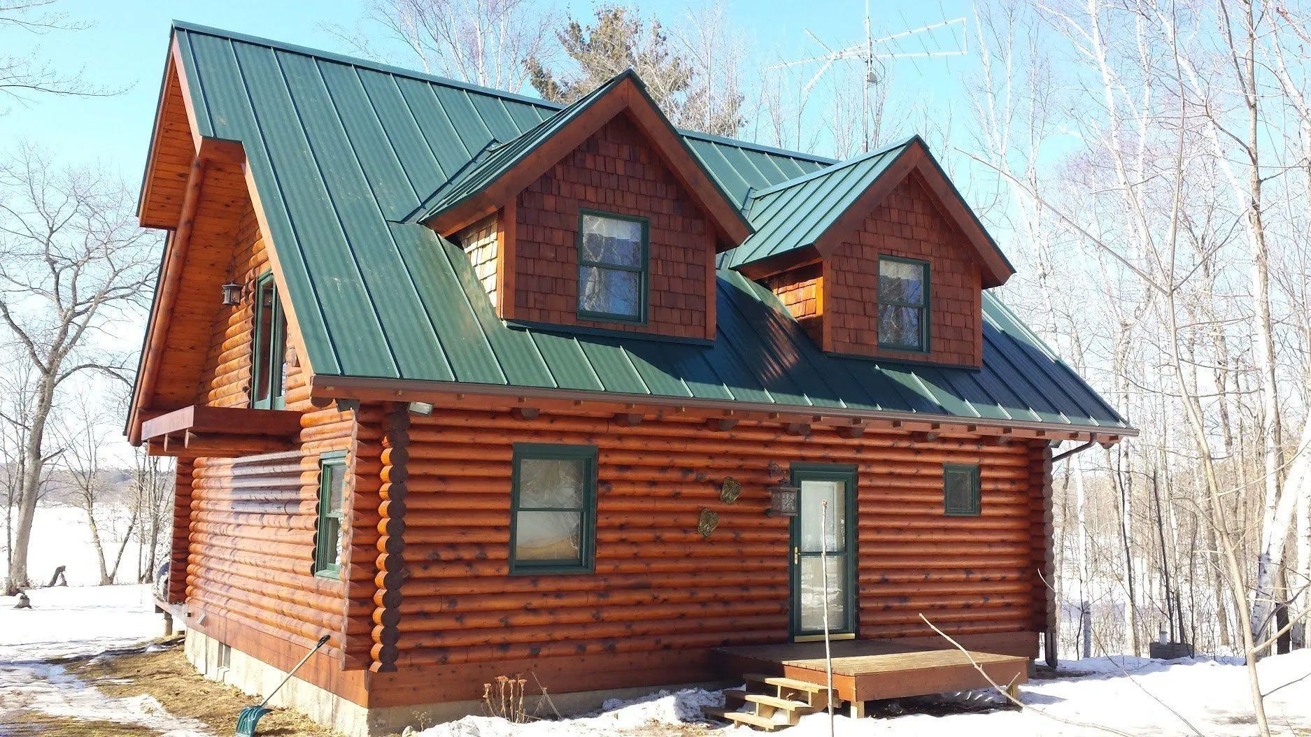 Snowy log cabin with green metal roof and dormer windows among bare trees
