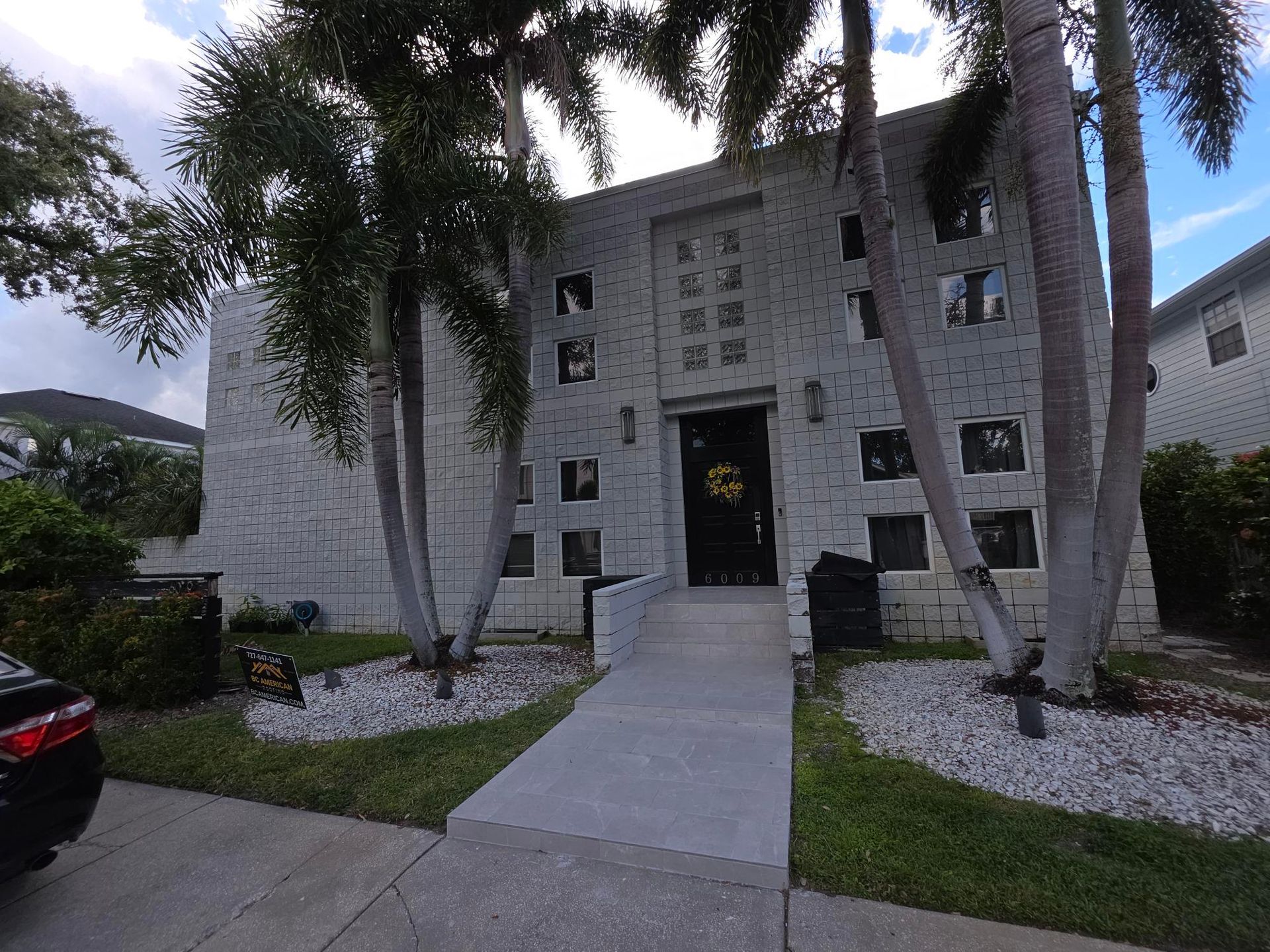 Two-story light-colored brick building with palm trees in front; concrete path leading to a dark doorway.