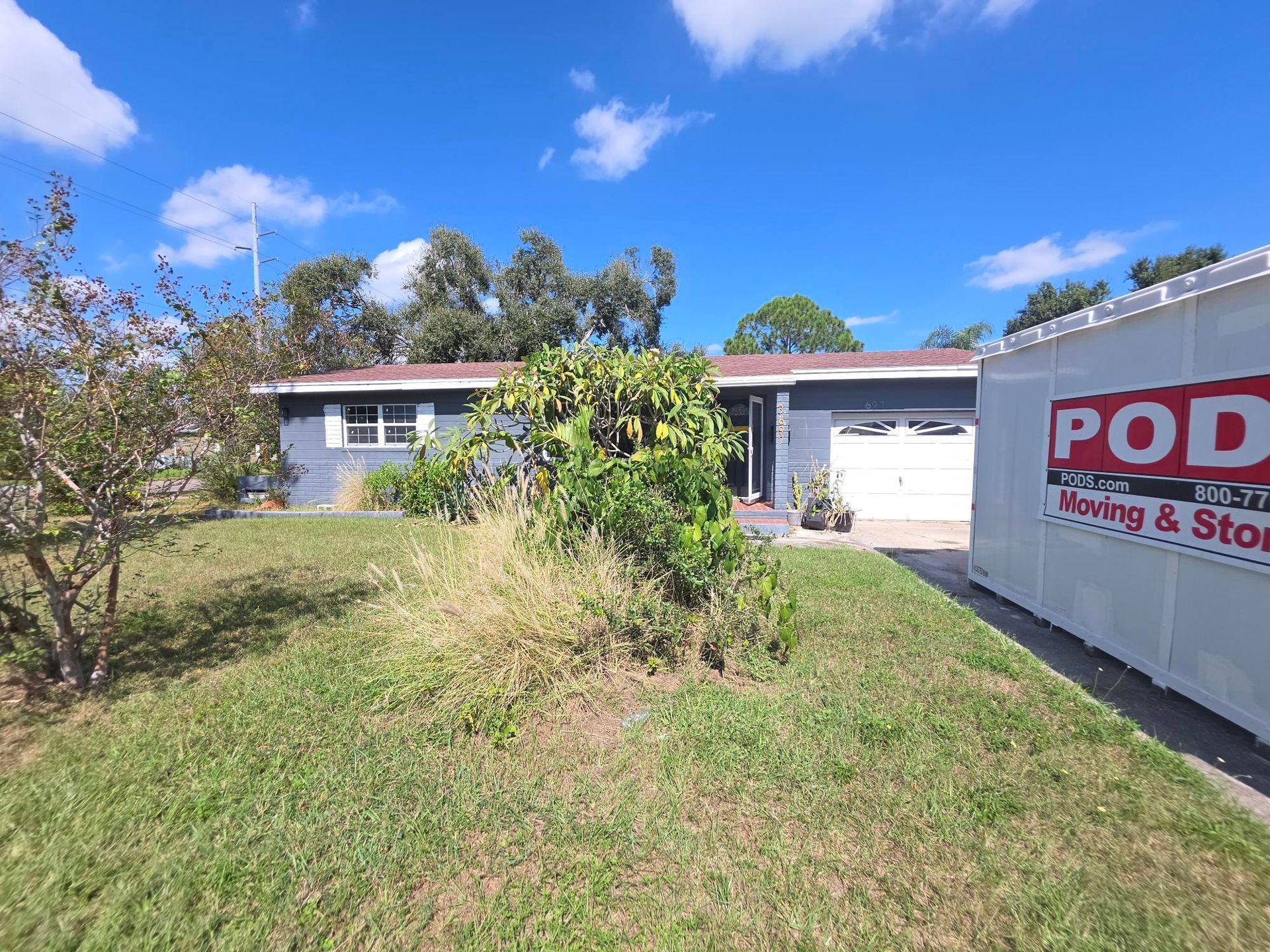 Blue house with white trim, green lawn, and a PODS container under a bright blue sky.