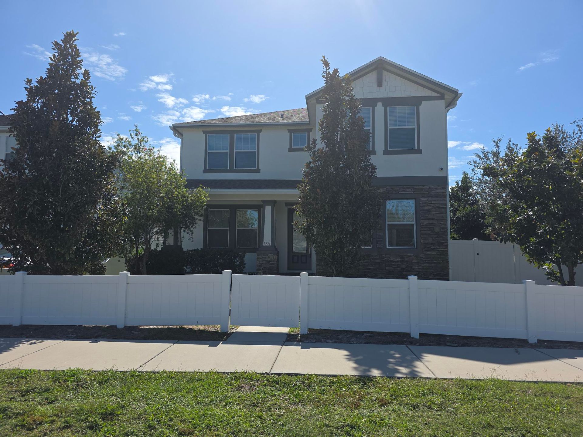 Two-story house with a white fence, blue sky, and trees.