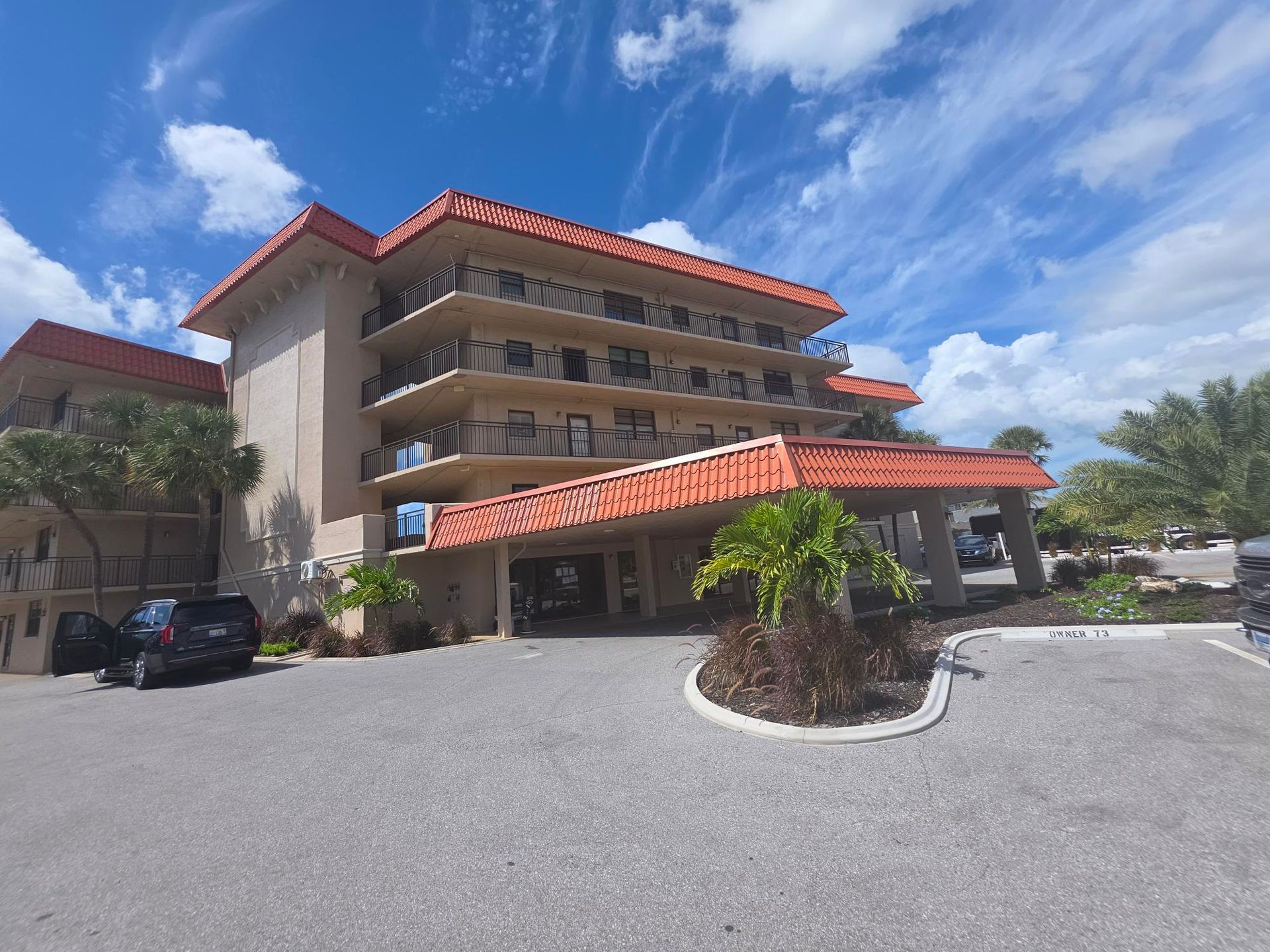 A beige multi-story building with red tile roof, balconies, and a covered entrance on a sunny day.