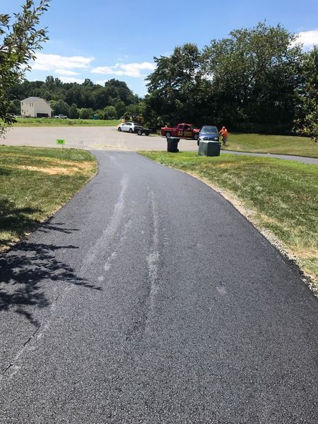 Curved asphalt path leading to a parking lot with trash bins and trees on a sunny day