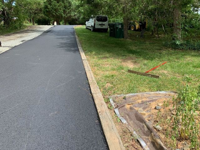 Freshly paved road beside grassy lot with a parked white van and green bins under trees