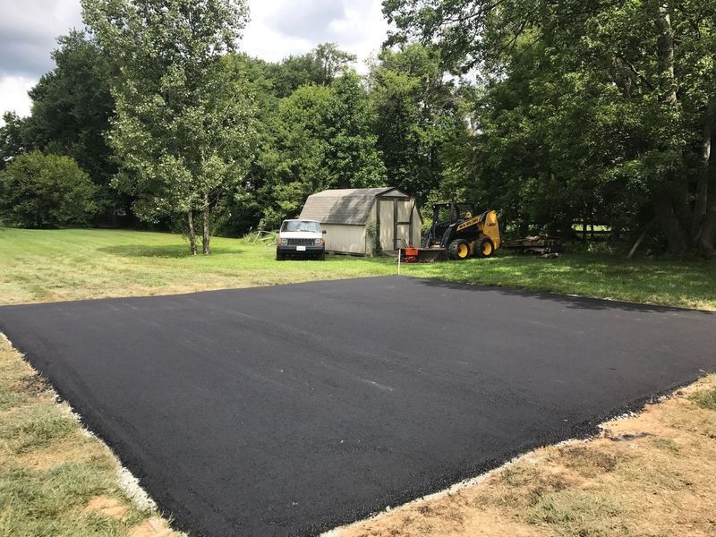 Fresh black asphalt driveway in a wooded yard with a car, shed, and backhoe nearby