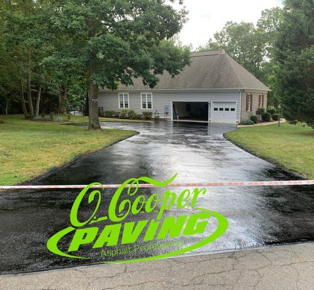 Freshly sealed black driveway leading to a garage, with a green “Cooper Paving” logo in the foreground.