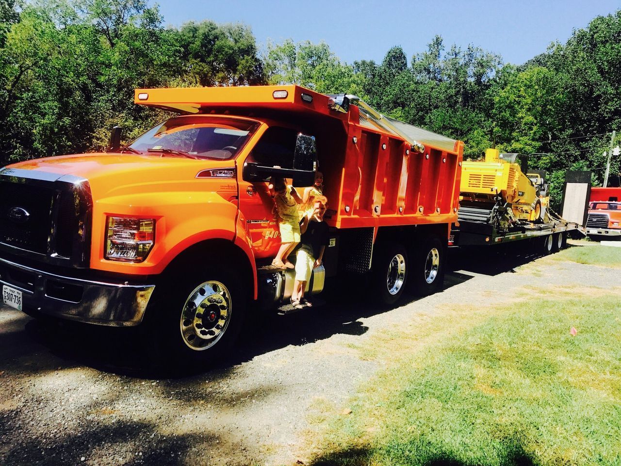 Orange dump truck towing yellow equipment on a road beside a grassy area and trees