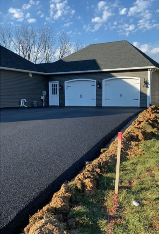 New asphalt driveway leads to a gray house with a two-car garage under a partly cloudy sky.