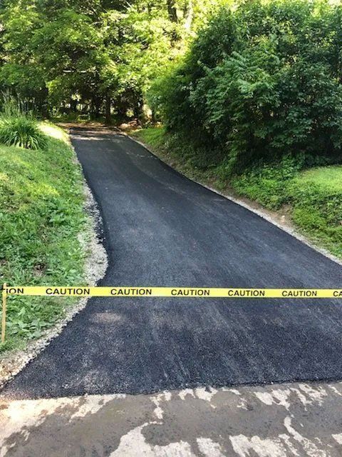Newly paved asphalt driveway blocked by yellow caution tape. Trees line the sides.
