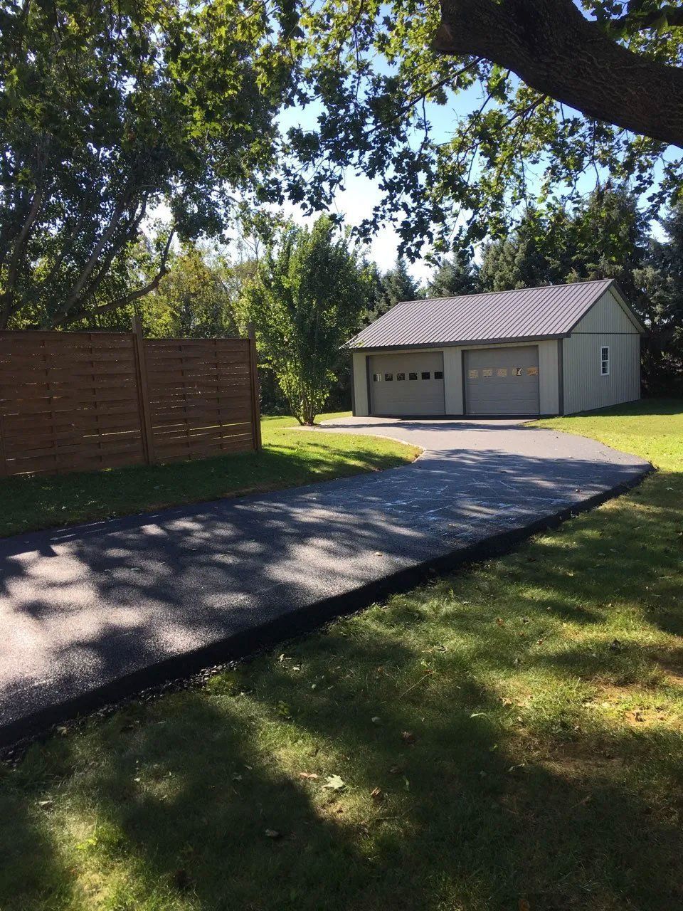 Asphalt driveway leading to a two-car garage with gray doors; brown fence on the left; trees.
