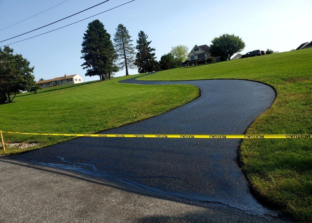 Newly paved black asphalt driveway winding uphill through green grass. Yellow caution tape in foreground.