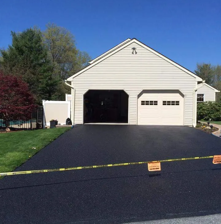 Freshly paved black driveway in front of a light-colored two-car garage under a clear blue sky.