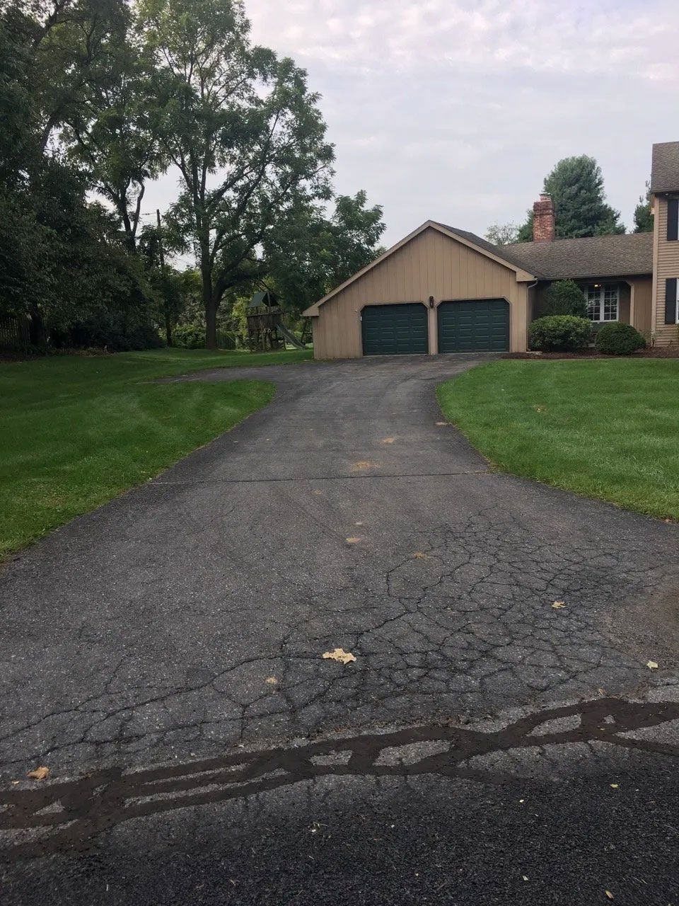 Asphalt driveway leading to a tan house with green garage doors; green lawn surrounds.