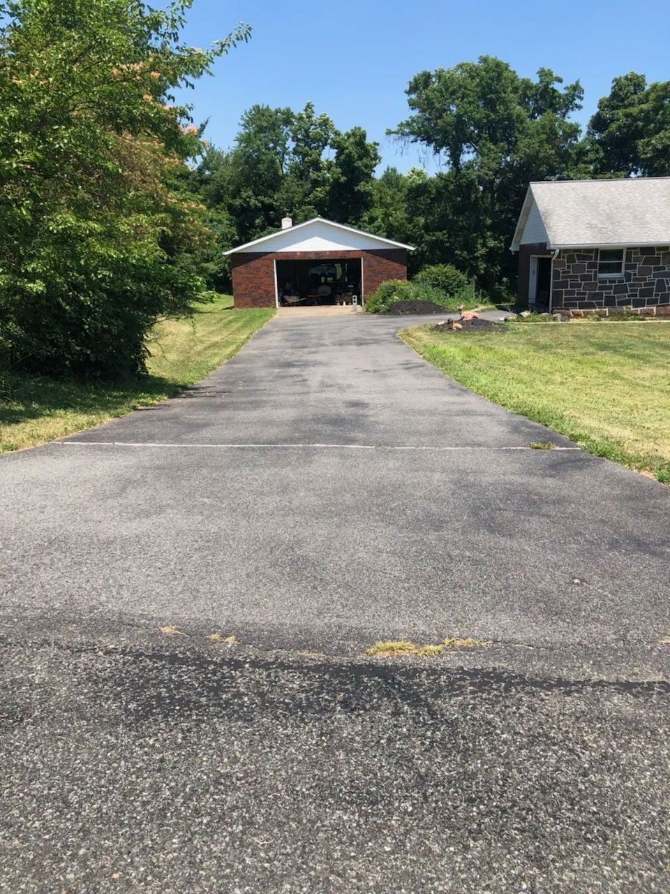 Driveway leading to an open garage; grass on either side. A stone house is to the right. Bright, sunny day.