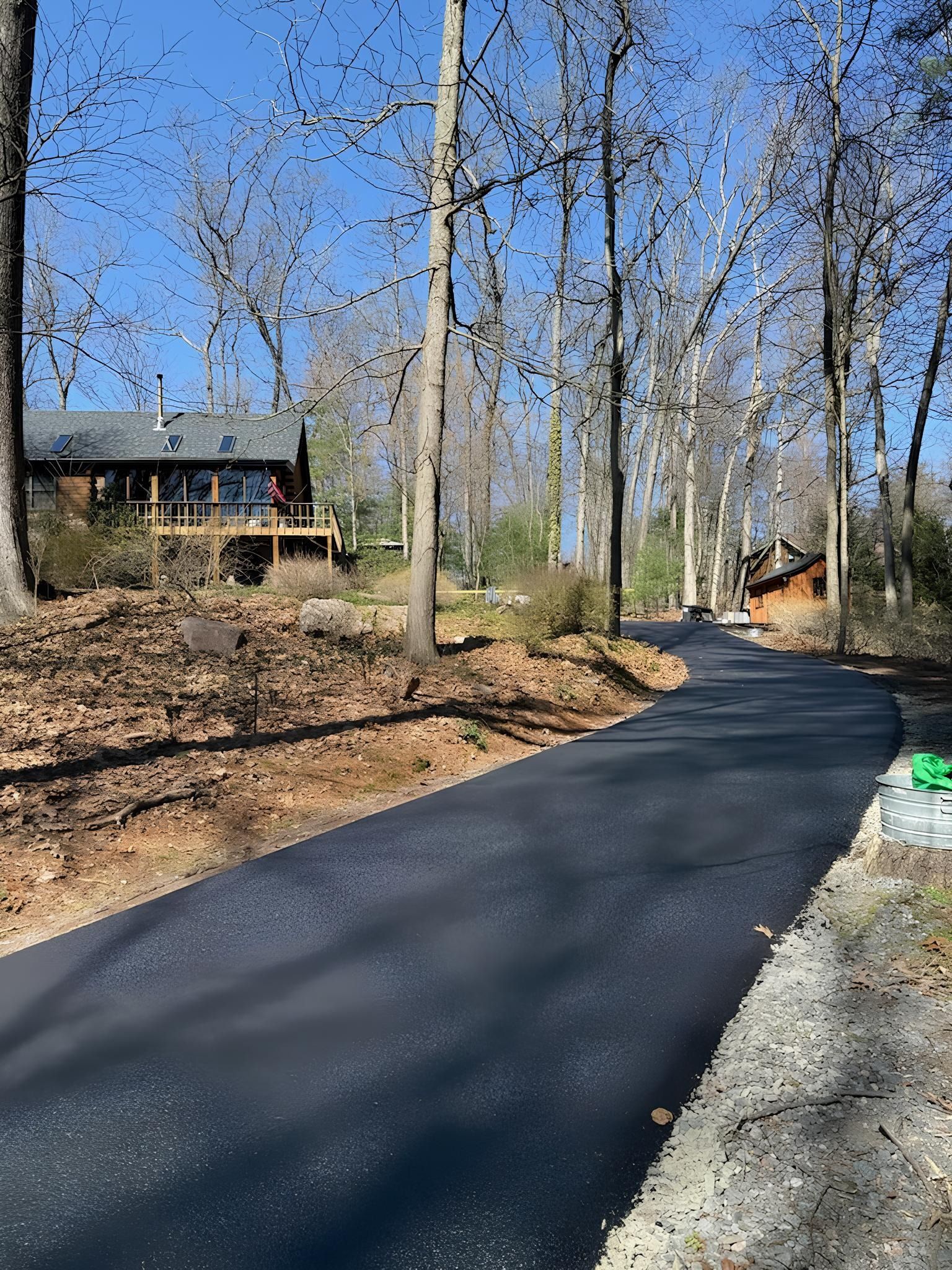 A freshly paved asphalt driveway winds through a wooded area toward a rustic house in the background.
