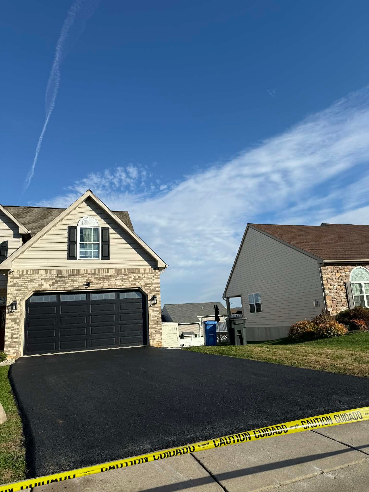 Newly paved black asphalt driveway in front of a tan brick house with a dark garage door, and a brown roof.