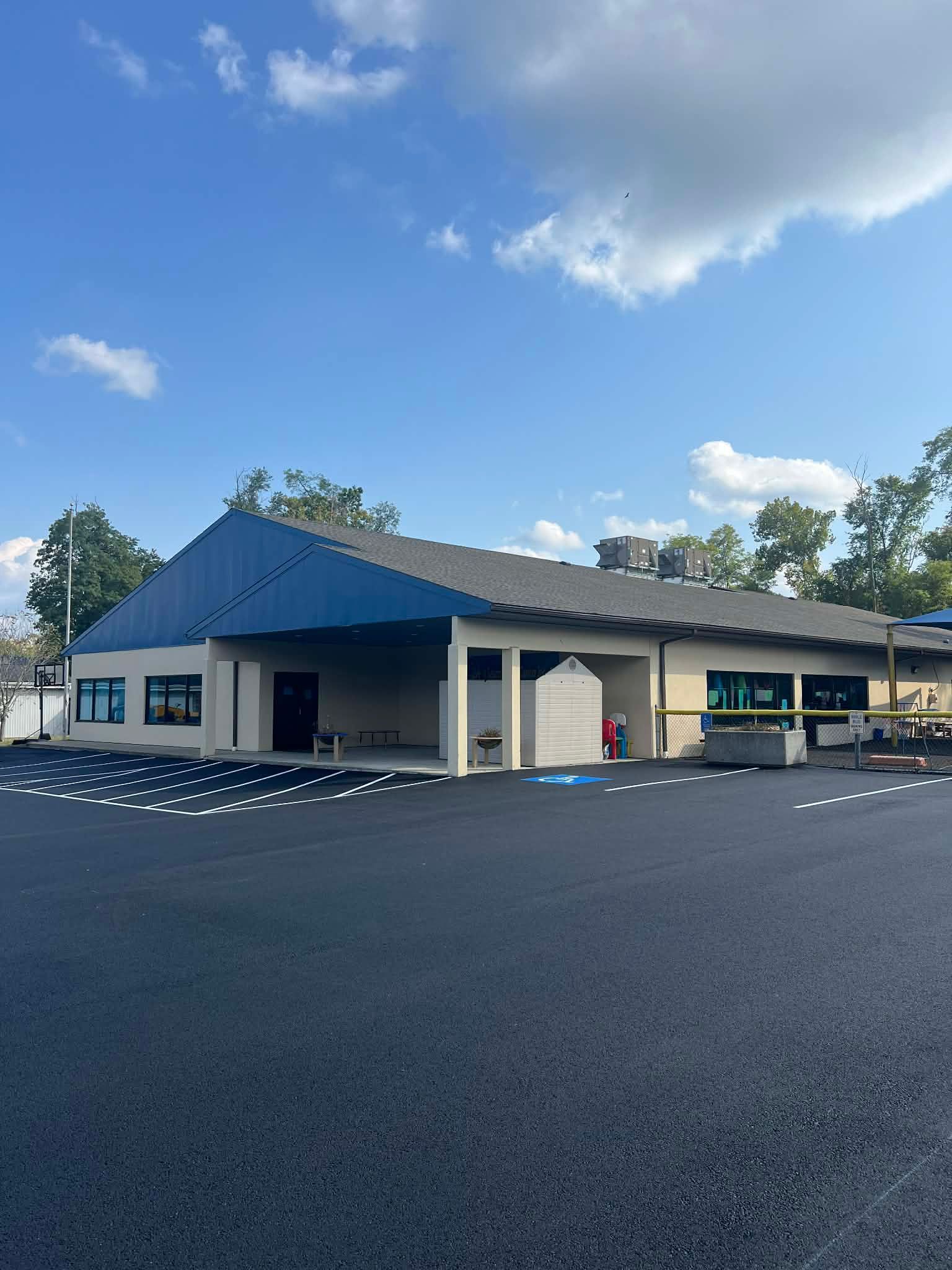 Exterior view of a building with a blue roof and beige walls. Asphalt parking lot in front. Sunny day.