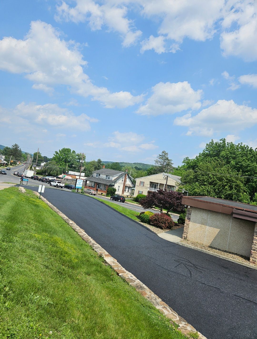 Newly paved road slopes downhill past houses and green lawns under a blue sky with fluffy clouds.