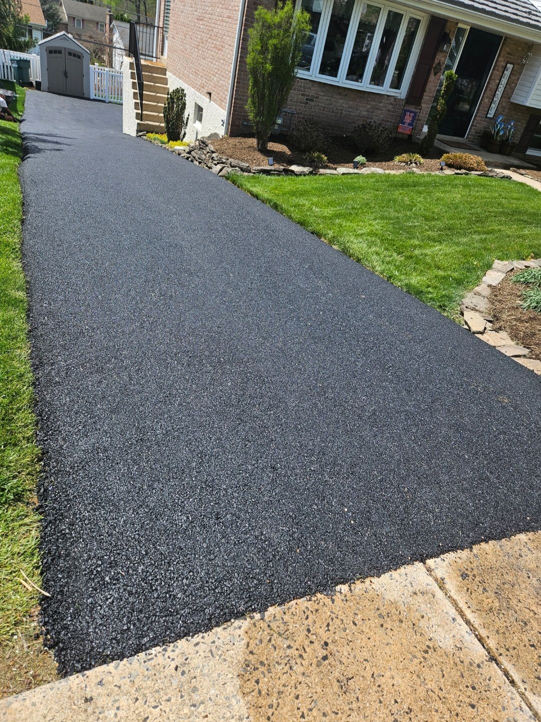 Newly paved asphalt driveway leading to a house, flanked by green grass and sidewalk.