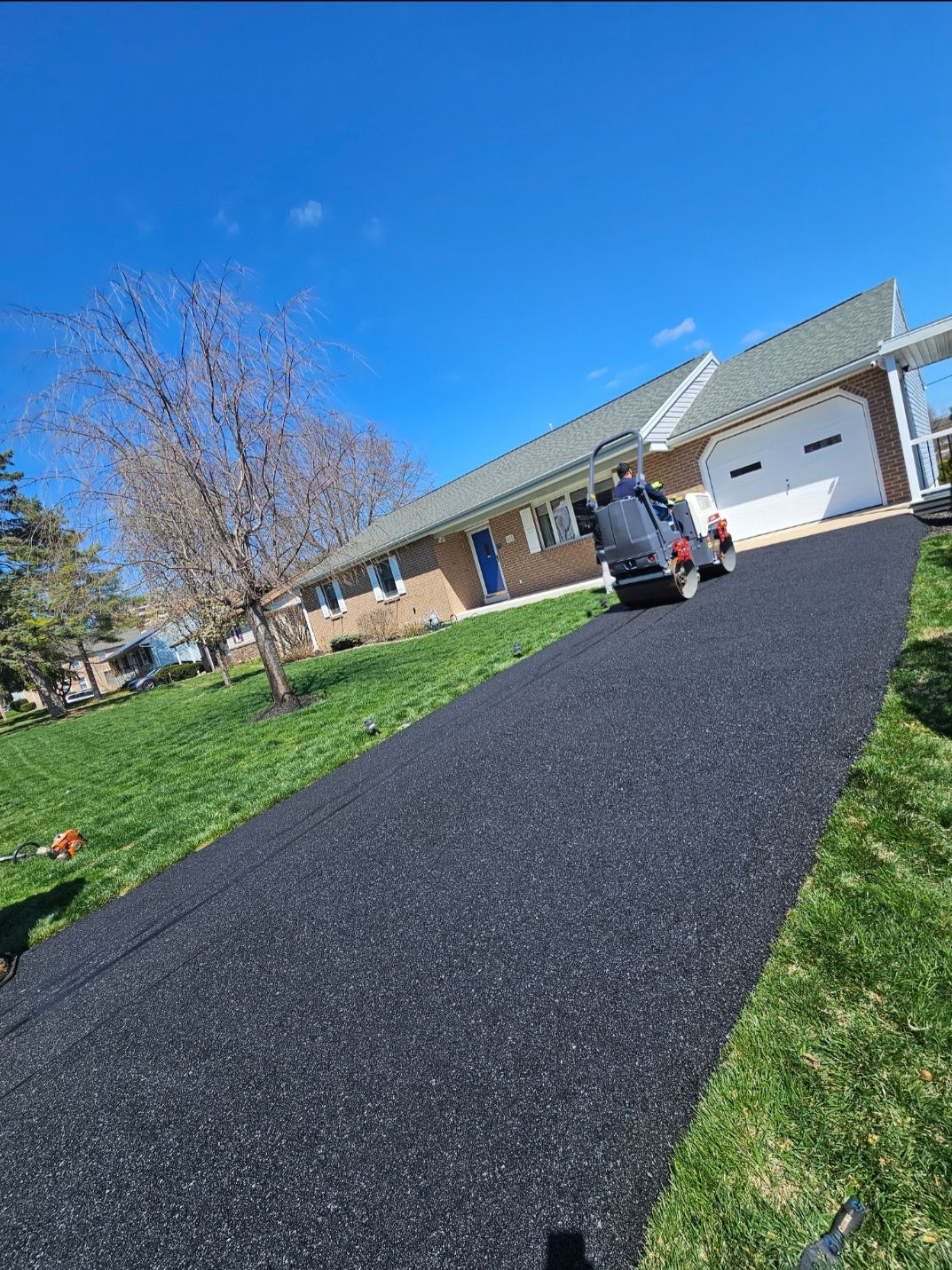 Newly paved black driveway leading to a brick house with a riding lawnmower on it. Sunny day, blue sky.