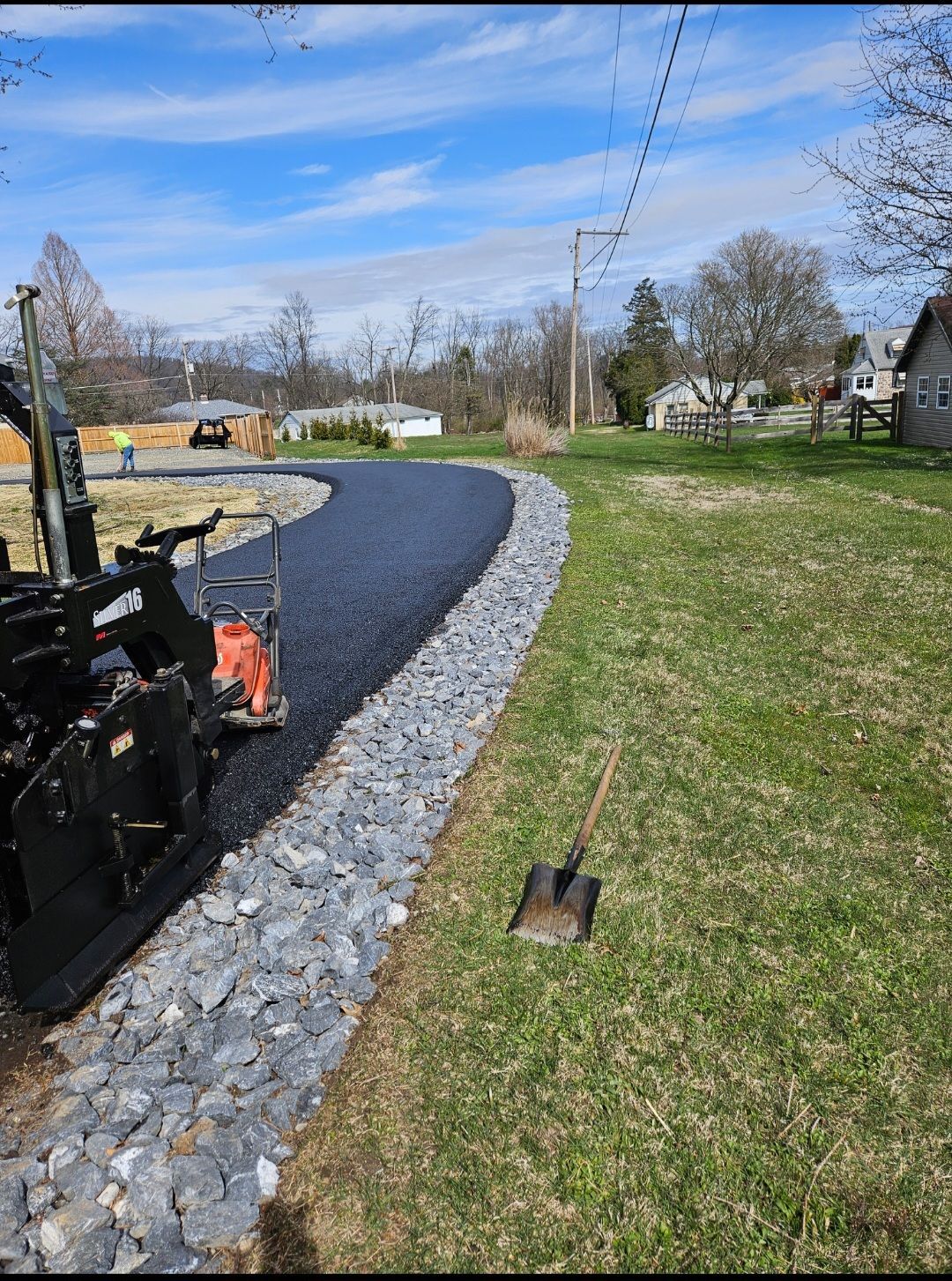 Asphalt paving along a rock-lined edge, with a shovel in the grass. A roller machine is in use, trees, and houses in the background.