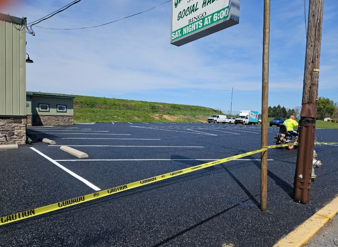 Newly paved parking lot with yellow caution tape blocking access; building and hill in the background.