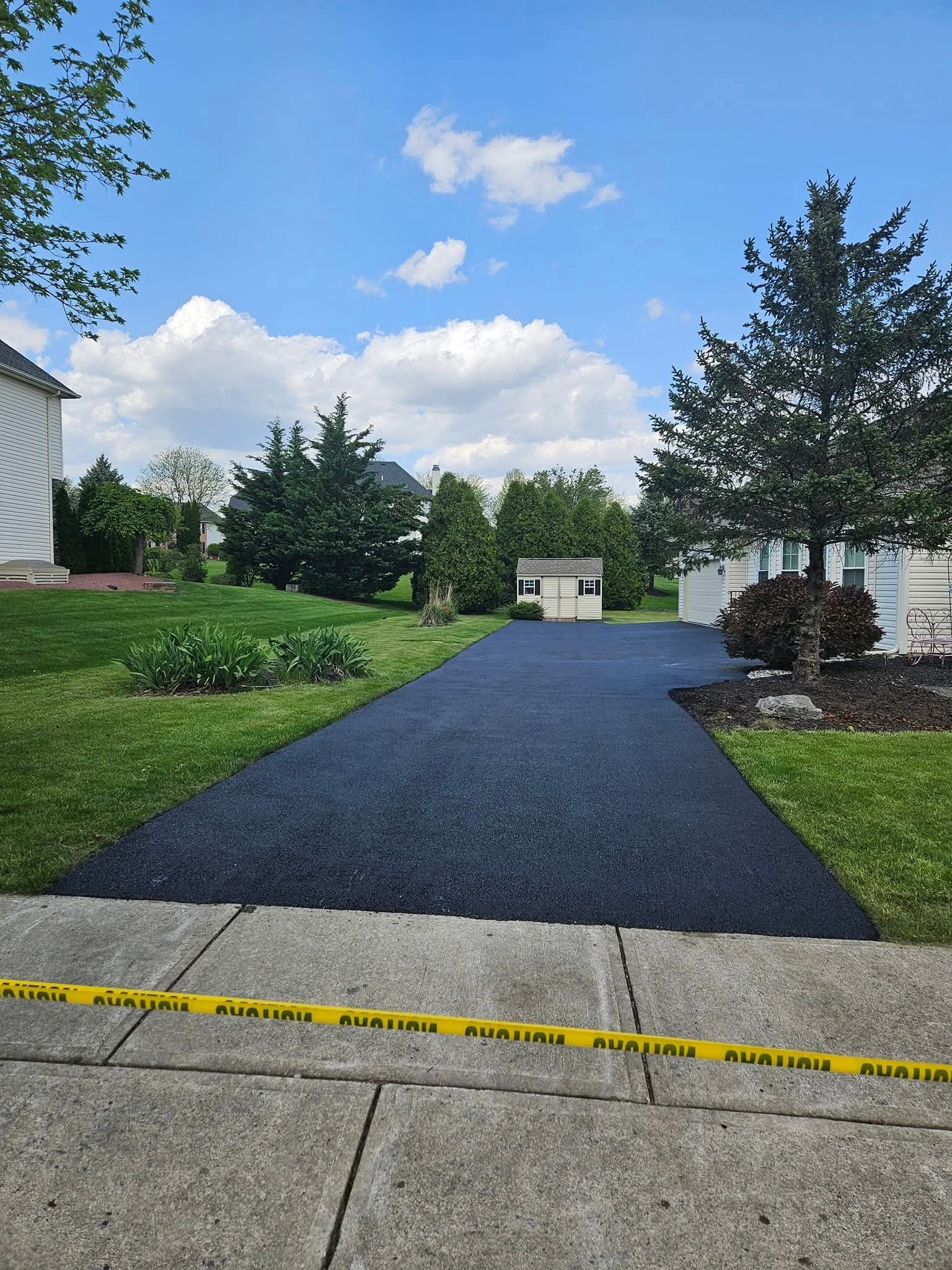 Freshly paved black driveway bordered by green grass and a sidewalk under a cloudy sky.