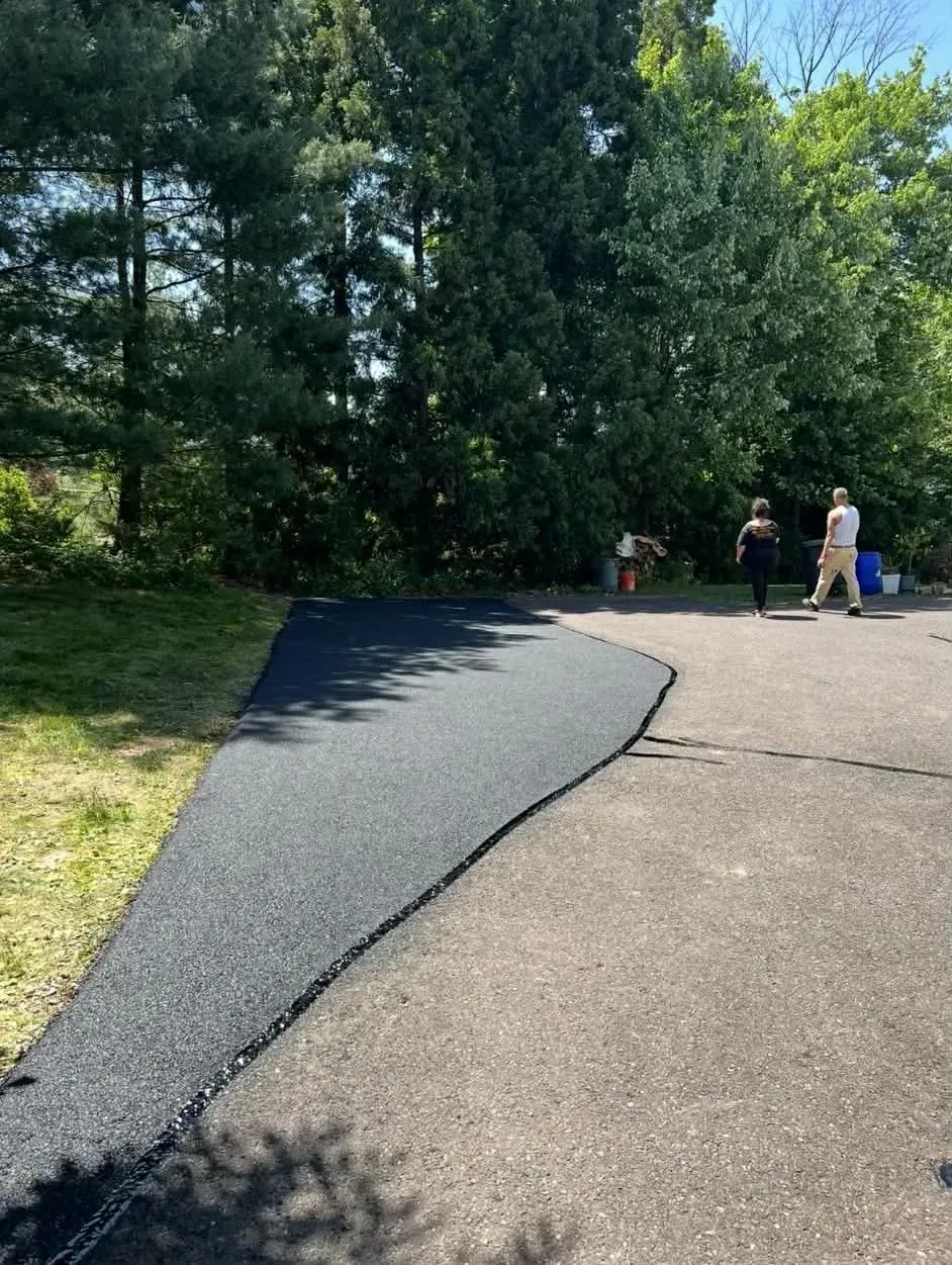 Two people walk on an old asphalt driveway next to a newly paved, dark black section extending onto the grass.