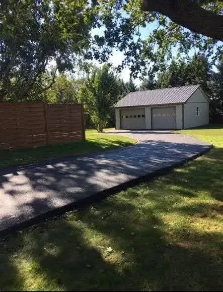 A paved driveway leads to a light-colored detached garage near a wooden fence and trees on a sunny day.