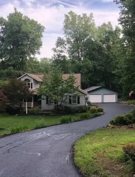 A two-story house with a detached two-car garage at the end of a long, paved driveway surrounded by trees and greenery.