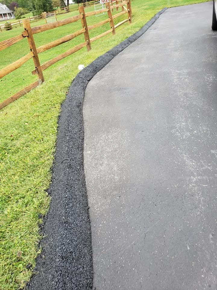 A freshly applied asphalt berm along the edge of a paved driveway next to a wooden fence and green grass.