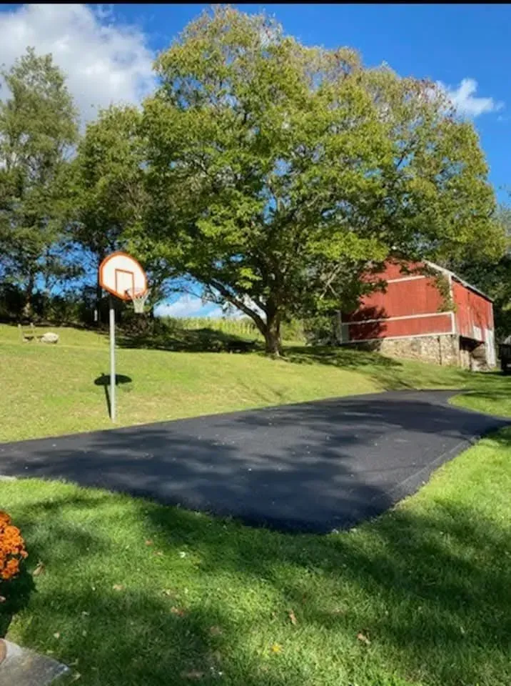 A basketball hoop stands on a paved driveway next to a large tree and a rustic red barn on a grassy lawn.