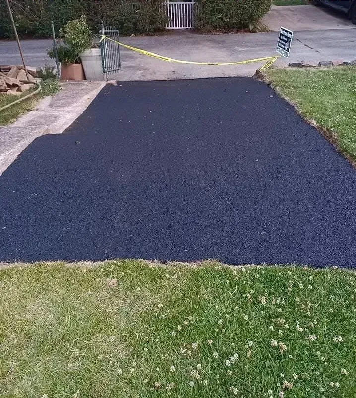 A freshly paved asphalt driveway section surrounded by green grass and a yellow caution tape boundary.