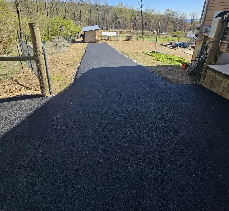 A freshly paved asphalt driveway leading from a structure toward a shed in a rural, sunny outdoor setting.