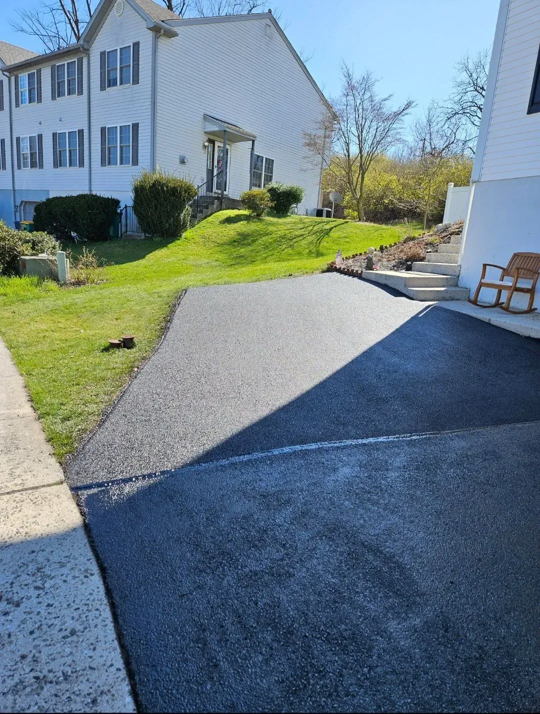 A freshly paved asphalt driveway sits in front of a white residential building with a green grassy lawn and stairs.